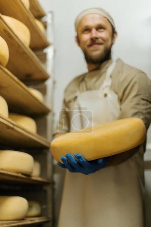 Photo for Cheese maker in uniform at cheese production man stands in warehouse with wooden shelves full cheese heads holds cheese in hand - Royalty Free Image