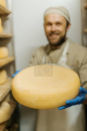 Foto de Fabricante de queso en uniforme en la producción de queso hombre se encuentra en el almacén con estantes de madera cabezas llenas de queso tiene queso en la mano - Imagen libre de derechos