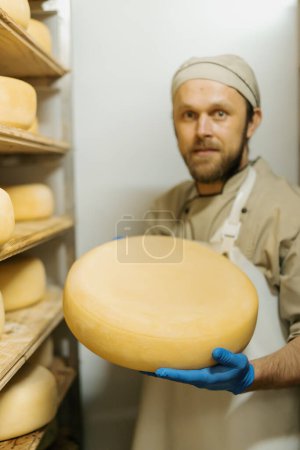 Foto de Fabricante de queso en uniforme en la producción de queso hombre se encuentra en el almacén con estantes de madera cabezas llenas de queso tiene queso en la mano - Imagen libre de derechos
