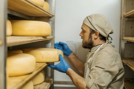 Foto de Fabricante de queso en uniforme en el hombre de producción de queso en el almacén con estantes de madera cabezas llenas de queso toma queso - Imagen libre de derechos