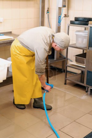 Photo for A worker at the production of a cheese factory washes the floor with water from a hose process of cleaning the workplace - Royalty Free Image