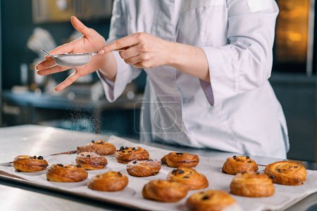 Foto de Atractivo panadero espolvorea azúcar en polvo listo recién horneado panecillos fragantes calientes panadería de producción de hornear - Imagen libre de derechos