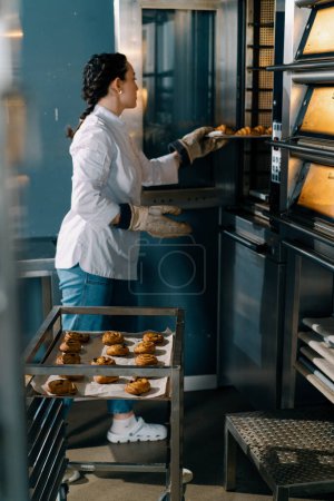 Photo for Attractive baker takes ready buns and croissants professional oven bakery production fresh baked goods - Royalty Free Image