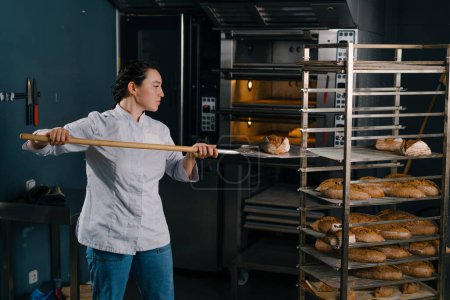 Foto de Panadero femenino saca pan fresco recién horneado del horno y lo pone en el estante en la cocina de la panadería profesión culinaria - Imagen libre de derechos