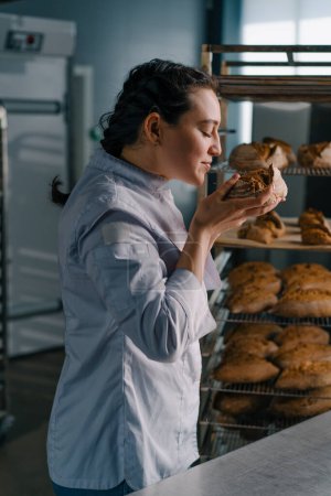 Photo for Attractive female baker takes freshly baked fragrant bread from the shelf sniffs it checks quality bakery professional kitchen baking - Royalty Free Image