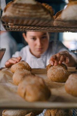Photo for Attractive female baker between shelves looking and checking freshly baked bread very carefully bakery industry - Royalty Free Image