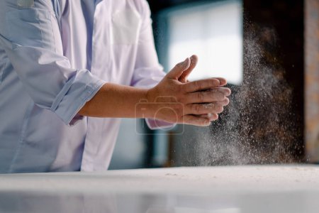Foto de Un chef experimentado se aplica harina en las manos antes de hacer pan en una panadería prepara pasteles en la cocina profesional - Imagen libre de derechos