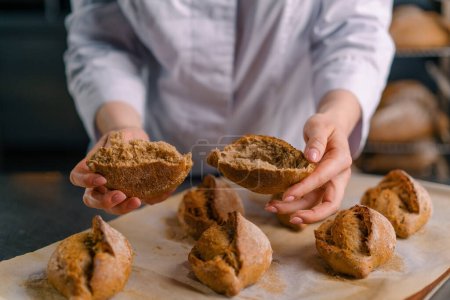 Foto de El panadero rasga el pan crujiente y esponjoso recién horneado con sus manos el bollo la panadería la producción pasteles - Imagen libre de derechos