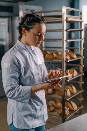 Téléchargez les photos : Une femme boulangère en uniforme près des étagères avec du pain fraîchement cuit tient la tablette dans ses mains vérifie la boulangerie de pain - en image libre de droit
