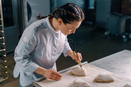 Photo for Beautiful woman baker cuts patterns with a professional baker's knife on raw bread buns before baking bakery production pastries - Royalty Free Image