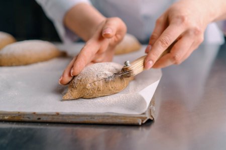 Photo for Woman baker cuts patterns on raw bread rolls with a professional baker's knife before baking bakery production pastries close-up - Royalty Free Image
