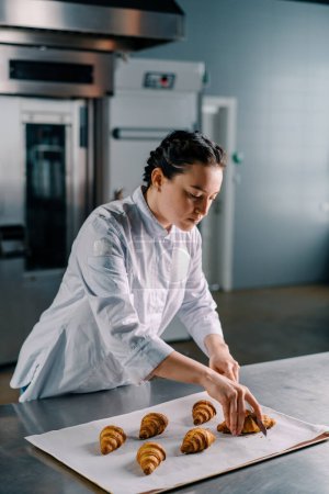 Photo for Beautiful woman baker cuts ready freshly baked hot crispy aromatic croissants with a knife and checks dough bakery production - Royalty Free Image