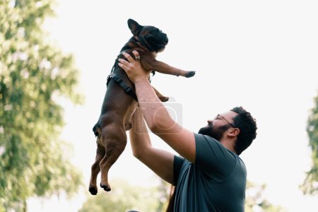 Foto de Barbudo hombre levanta su perro bulldog francés al cielo en el parque jugando y acariciando lindo perrito - Imagen libre de derechos