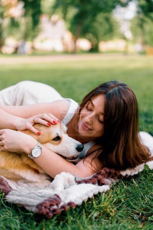 Photo for Young beautiful girl resting in nature lying on a sheet in the park stroking and kissing small corgi dog animals in nature - Royalty Free Image