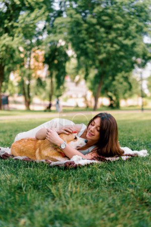 Foto de Joven hermosa chica descansando en la naturaleza acostado en una sábana en el parque acariciando y besando pequeños corgi perro animales en la naturaleza - Imagen libre de derechos