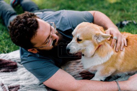 Téléchargez les photos : Homme barbu reposant dans le parc avec son chien corgi animal de compagnie à l'extérieur couché sur la feuille pique-nique d'été - en image libre de droit