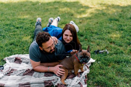 Photo for A married couple rests in nature lies on a sheet in the park plays with a small dog of French bulldog breed - Royalty Free Image