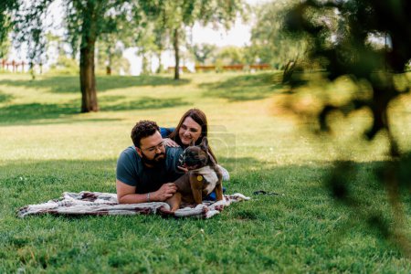 Téléchargez les photos : Un couple marié repose dans la nature repose sur un drap dans le parc joue avec un petit chien de race bouledogue française - en image libre de droit