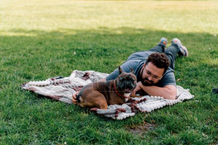 Photo for Bearded man resting in the park with his pet dog french bulldog outdoors lying on sheet summer picnic - Royalty Free Image