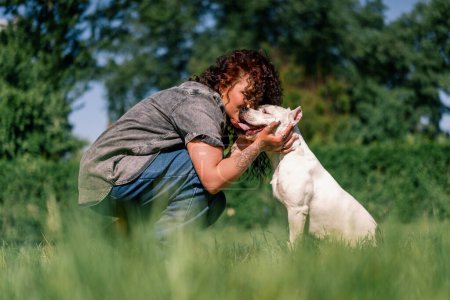 Foto de Psicólogo perro calma a un perro blanco de una raza de pit bull grande en el parque una mascota con una lengua sobresaliente estresado - Imagen libre de derechos