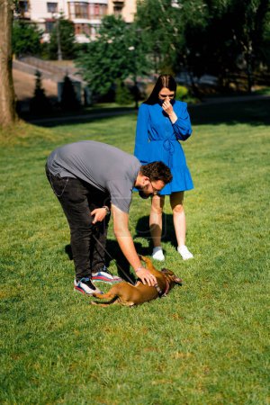 Foto de Una pareja familiar en un paseo con un bulldog francés en el parque los propietarios están jugando con lindo perrito - Imagen libre de derechos