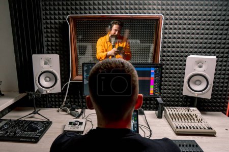 Photo for Young pop rap artist emotionally recording a new song in a professional recording studio sound engineer watching tracks on display - Royalty Free Image