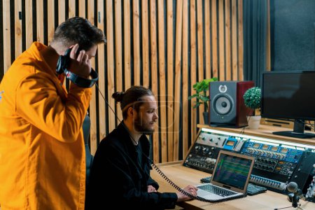 Téléchargez les photos : Jeune rappeur pop enregistrant émotionnellement une nouvelle chanson dans un studio d'enregistrement professionnel ingénieur du son regarder des pistes exposées - en image libre de droit