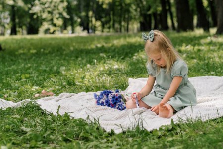 Photo for Portrait Cute little girl in a dress in the park draws a picture of a postcard with a felt-tip pen for congratulating parents with holiday - Royalty Free Image