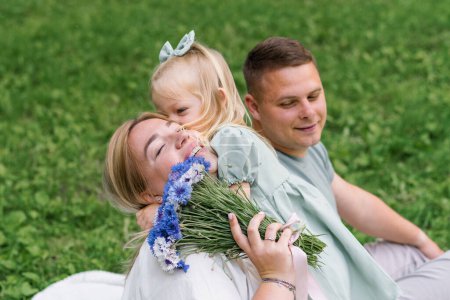 Foto de Una familia feliz descansa en la naturaleza en el parque, una pequeña hija en un vestido le da a su madre un ramo de flores padres felices besan a su hija - Imagen libre de derechos
