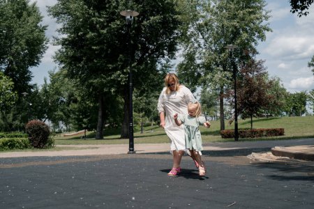 Photo for Little cute girl in a green summer dress with a bow on her head on the playground with her mother on walk holding hands child abduction - Royalty Free Image