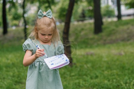 Téléchargez les photos : Portrait d'une fille capricieuse bouleversée en colère dans une robe dans le parc dessine une carte postale avec feutre stylo obtient ennuyé et esquisse tout ennuyeux - en image libre de droit