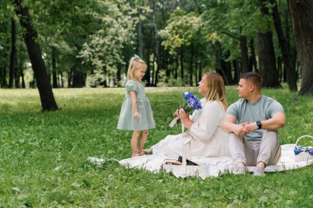 Foto de Una familia feliz descansa en la naturaleza en el parque una pequeña hija en un vestido da a su madre ramo de flores - Imagen libre de derechos