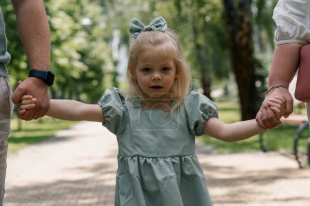 Photo for Close up portrait of cute little girl with bow walking in summer park holding hands of her parents Happy moments family weekend - Royalty Free Image