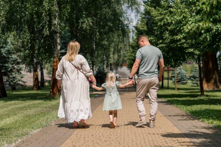 Photo for Little cute girl in green summer dress with parents in city park on walk summer family lifestyle Childhood happy family rear view - Royalty Free Image