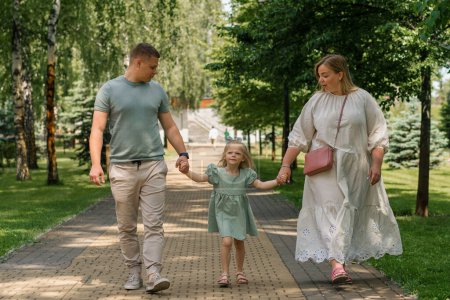 Téléchargez les photos : Petite fille mignonne en robe d'été verte avec les parents dans le parc de la ville sur la marche style de vie de famille d'été Enfance famille heureuse tenant la main - en image libre de droit