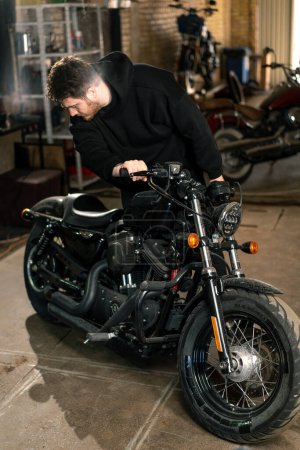 Foto de Serios motorista barbudo en sudadera con capucha negro en taller de motocicleta de pie cerca de la motocicleta inspeccionarlo para su reparación - Imagen libre de derechos