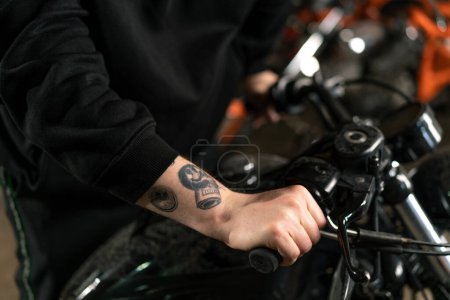 Foto de Primer plano de la mano del motociclista tatuado que sostiene el manillar de la motocicleta en el taller de control de garaje perilla de gas - Imagen libre de derechos