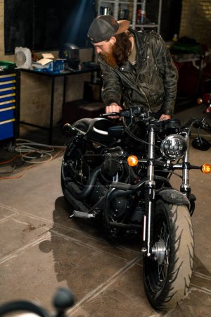 Foto de Taller creativo de motocicleta auténtica Garaje pelirroja barbuda mecánico de pie cerca de la motocicleta comprobarlo - Imagen libre de derechos