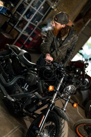 Photo for Creative authentic motorcycle workshop Garage redhead bearded biker mechanic smoking cigarette near motorcycle - Royalty Free Image