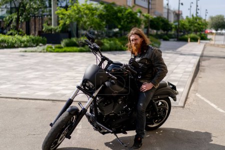 Photo for Brutal red-haired biker with a beard sits on a black motorcycle before the start of the race holding a helmet in his hands thirst for speed - Royalty Free Image
