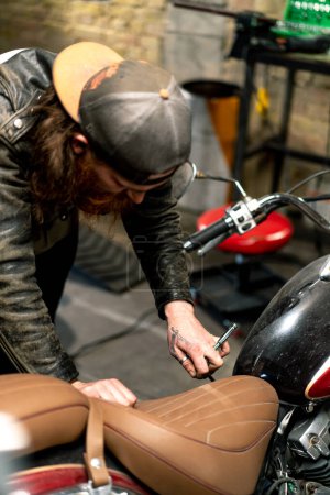 Téléchargez les photos : Atelier de Moto Authentique Créatif Garage Rouge Barbu Biker Mécanicien Concentré Réparation Siège de Moto - en image libre de droit