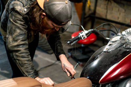 Foto de Taller creativo de motocicleta auténtica Garaje rojo barbudo motorista mecánico concentrado reparación asiento de la motocicleta - Imagen libre de derechos