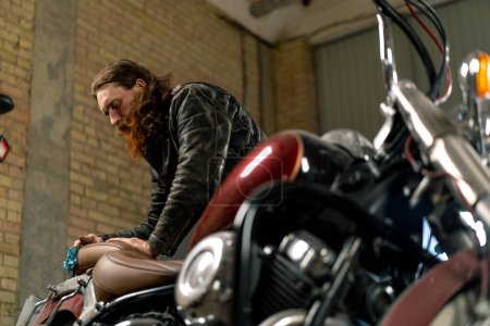 Téléchargez les photos : Atelier de moto authentique créatif Garage de motard barbu rouge mécanicien essuyant belle moto avec chiffon - en image libre de droit
