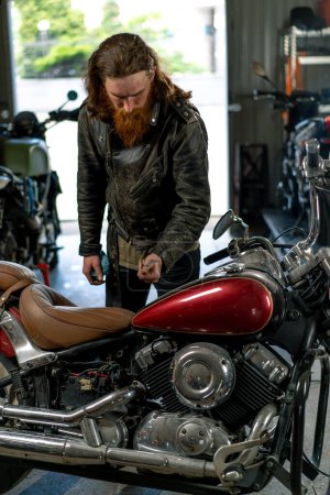Photo for Creative authentic motorcycle workshop garage redhead bearded biker mechanic repairing and looking at beautiful motorcycle - Royalty Free Image