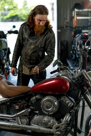 Photo for Creative authentic motorcycle workshop garage redhead bearded biker mechanic repairing and looking at beautiful motorcycle - Royalty Free Image