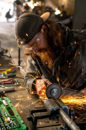 Photo for Creative authentic motorcycle workshop redhead bearded brutal biker works with circular saw Sparks fly from hot metal - Royalty Free Image