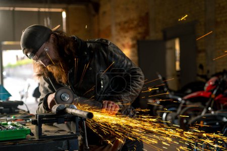 Photo for Creative authentic motorcycle workshop redhead bearded brutal biker works with circular saw Sparks fly from hot metal - Royalty Free Image