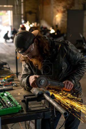 Téléchargez les photos : Atelier de moto créatif authentique rousse barbu motard brutal travaille avec scie circulaire Sparks voler de métal chaud - en image libre de droit