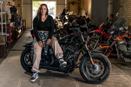Foto de Creativo taller de motocicleta auténtica garaje hermosa joven ciclista sentado en una motocicleta fresca - Imagen libre de derechos