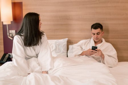 Foto de Retrato de una joven pareja en su luna de miel en una habitación de hotel acostada en la cama con túnicas blancas discutiendo el descontento de conflicto - Imagen libre de derechos